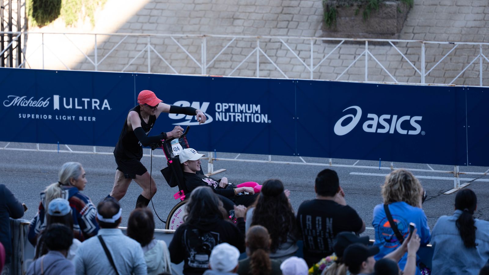 A runner pushes a participant in a racing chair past spectators near the finish line of the Los Angeles Marathon on Sunday, March 8, 2026, in Los Angeles.